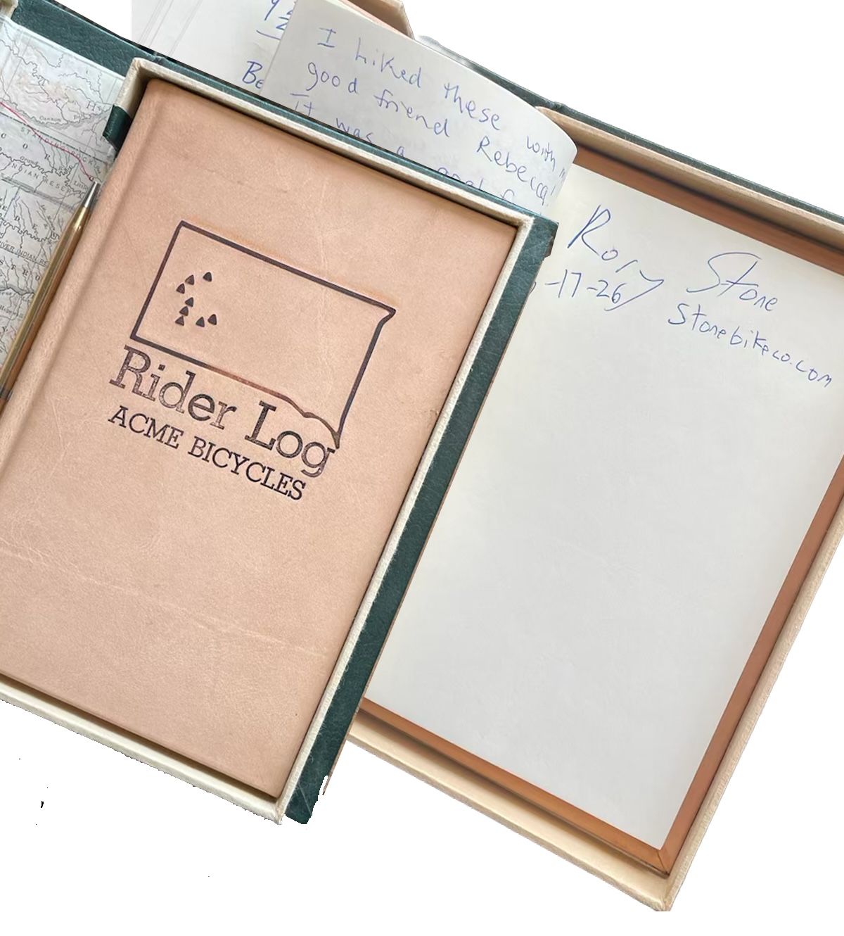 The Acme Bicycles Rider Log — leather-bound journal stamped with the South Dakota state outline and the 8/7 summit cluster, open to a signed finisher page