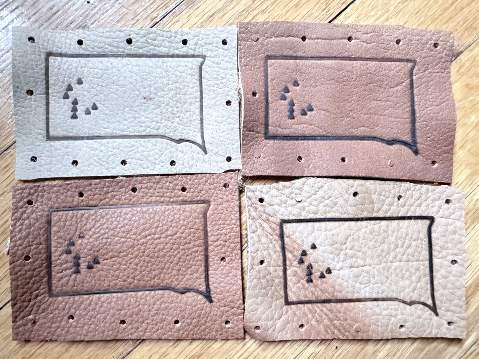 Four hand-cut leather 8 Over 7 finisher patches arranged in a grid — the South Dakota state outline framing the eight summit triangles, stitch-holes punched along every edge, four different leather tones from oyster to chestnut